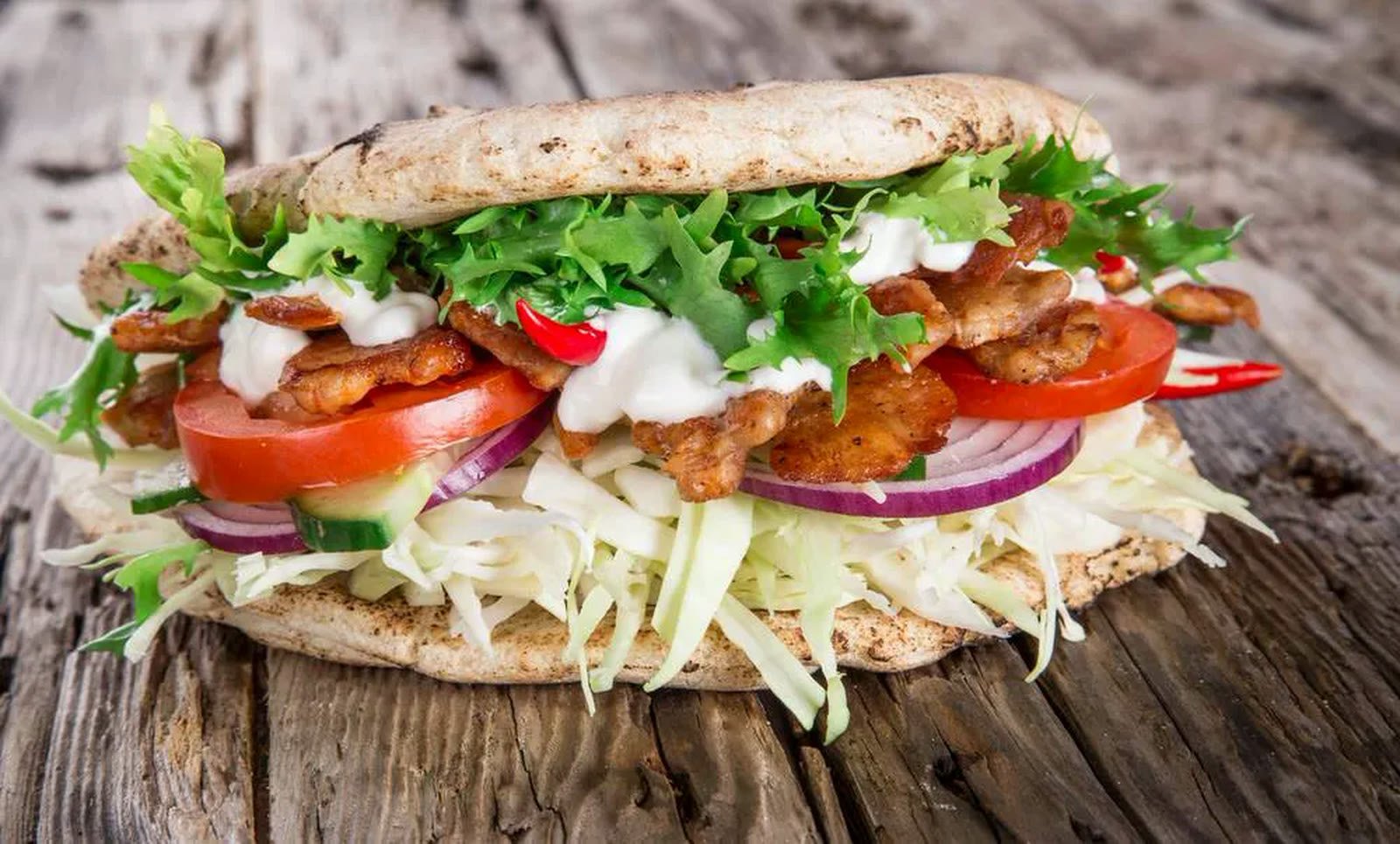 Een broodje met gegrild vlees, sla, tomaat en witte saus op een houten plank.
